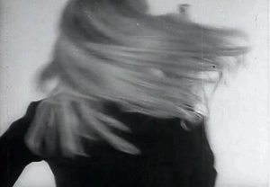 Nico's Screen Test: animated gif, Black-and-white image of Nico turning. She wears a tuxedo and holds a bottle of spirits. Her long blonde hair spins with the turn