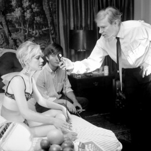 black-and-white photo on the set of Andy Warhol Beauty No. 2. Edie Sedgwick and Gino Piserchio on a bed as Andy Warhol holds a light meter in front of them.